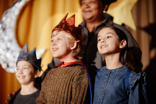 Three children wearing costumes standing on stage during school play in front of curtain. Smiling children in various costumes happily participating in event