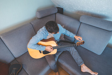Young man sits barefoot on a gray couch, carefully tuning his acoustic guitar. He wears a light...
