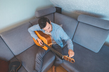 Young man sits barefoot on a gray couch, carefully tuning his acoustic guitar. He wears a light...