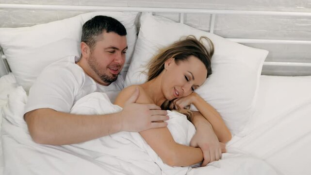 A young couple in love lie in bed over a blanket and hug. A man strokes a woman's shoulder and kisses her on the shoulder. Morning of Valentine's Day.