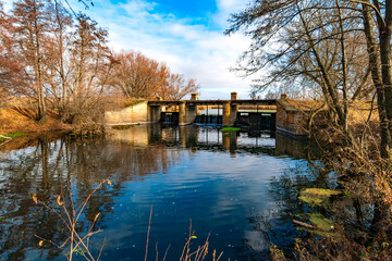 Obraz premium Historic dam structure over a calm river surrounded by bare autumn trees, reflecting the clear blue sky and tranquil waterscape