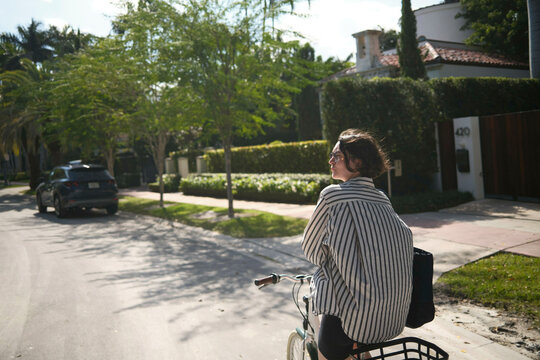Woman in striped shirt rides a bicycle on a sunny suburban street with lush greenery, Miami, Florida, USA