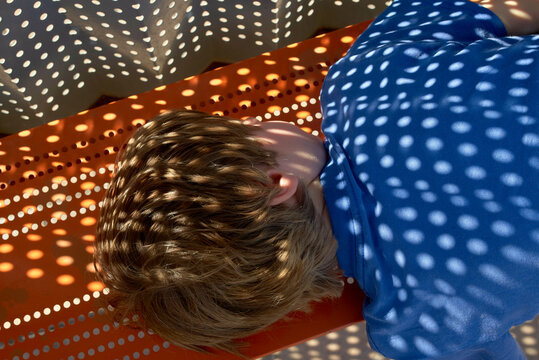 Child lying on an orange perforated bench, with light and shadow patterns on their head. Israel
