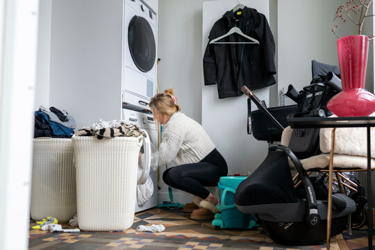 A woman kneels in front of a washing machine, loading laundry. The room is filled with household items,. Netherlands