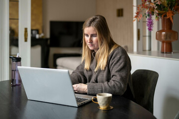 Woman with long blonde hair wearing a grey sweater works on a laptop at a black table, with a cup of coffee. Netherlands