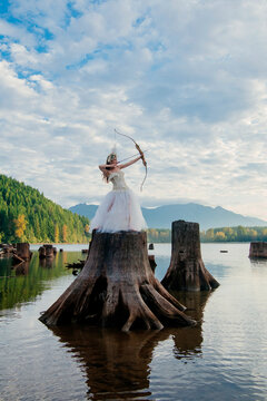 Woman in a white dress with a bow and arrow standing on a large tree stump in a lake. Rattlesnake Lake, Puget Sound, WA, USA