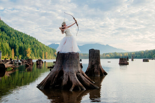 Woman in a white dress aiming a bow and arrow on a large tree stump in a serene lake.   Rattlesnake Lake, Puget Sound, WA, USA