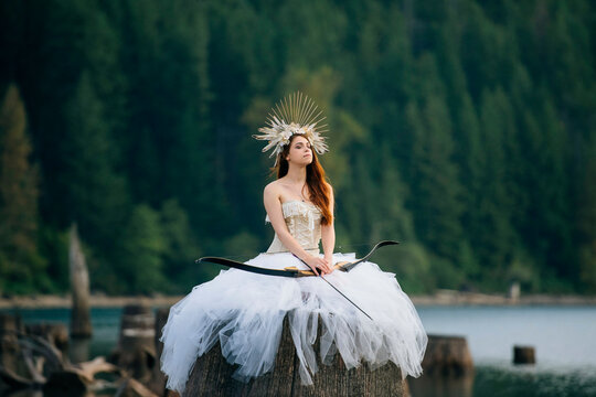 Woman in white gown with crown and bow and arrow sits on a stump by a lake and forest. Rattlesnake Lake, Puget Sound, WA, USA