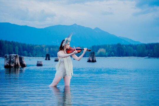 Woman playing violin in shallow lake surrounded by mountains and tree stumps. Rattlesnake Lake, Puget Sound, WA, USA