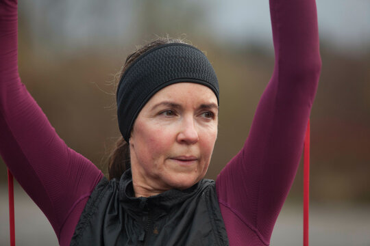 A focused adult woman stretching her arms above her head while exercising outdoors on a cloudy day. Sale Water Park, Manchester, UK