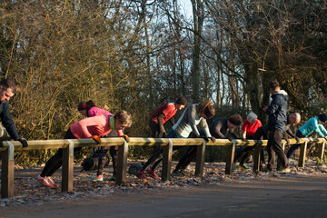 A group of people exercising outdoors, leaning against a wooden railing in a forested park area on a sunny day. Sale Water Park, Manchester, UK