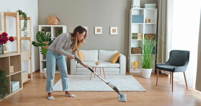 Smiling woman washing wooden floor with microfiber spray mop in bright living room