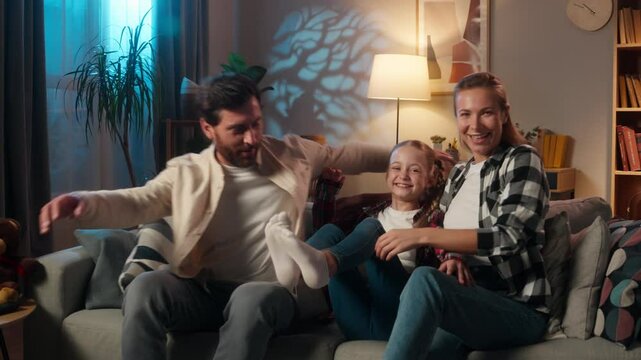 Caucasian family mother woman man father with child kid girl daughter together spending time relaxing laughing happy jumping falling on couch at home in evening night cozy sofa smiling loan mortgage