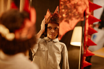 Young boy wearing dinosaur costume, adjusting red hat while looking in mirror during festive...