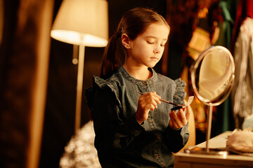 Young girl focusing on applying makeup while sitting in dimly lit room with mirror and lamp behind....