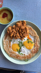 A plate of Roti Canai Sarang Burung, a popular Malaysian flatbread, topped with a savory chicken rendang and with half cooked fried eggs. A side of dip adds a touch of spice to this delicious dish