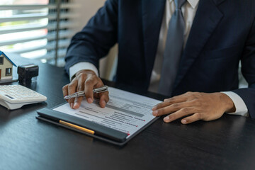 A man is sitting at a desk with a calculator and a piece of paper with numbers on it. He is pointing to the paper and he is explaining something to someone. The scene suggests a professional setting
