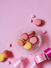 Colorful macarons on a glass plate and heart shape sugars with pink ribbons on a pink background. Valentine's day party table setting with a copy space
