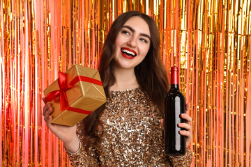 Happy young woman with gift box and bottle of wine against foil curtain