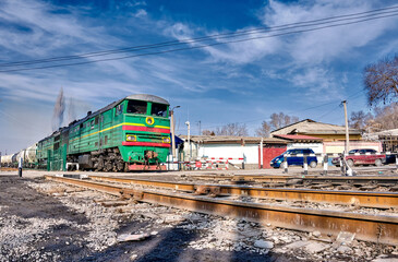A green locomotive pulls tank cars against a blue sky.