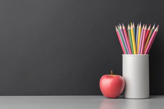 A colorful arrangement of pencils in a white cup alongside a shiny red apple against a dark background.