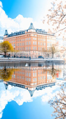 A picturesque building reflects in calm water under a clear blue sky, surrounded by light clouds and hints of autumn foliage.