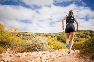 Fototapeta premium Female trail runner wearing black sportswear and a hydration vest, running through a scenic natural landscape on a rocky path.
