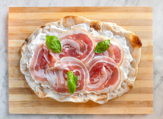 traditional pinsa romana stuffed with stracciatella fresh cheese, ham and basil hanging on a wooden cutting board