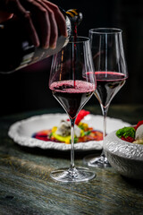 A close-up of red wine being poured into elegant glasses, accompanied by a gourmet dish on a decorative plate. The setting is warm and inviting, perfect for a fine dining experience.