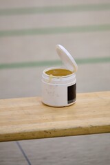 Open jar of sticky resin or adhesive with a partially closed lid sitting on a wooden bench indoors during a handball match.