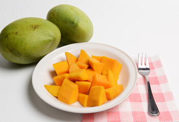 Cut mango fruit on a white plate and two fresh whole mangoes at the background.