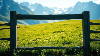 Fototapeta premium Rustic wooden fence framing a vibrant green meadow with a majestic mountain range in the background