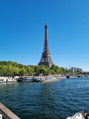 Fototapeta premium Scenic view of the Eiffel Tower in Paris, France, towering over the Seine River with tour boats docked along the banks, lush green trees lining the waterway, and a bright blue sky with a few clouds.