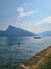 Obraz premium Serene lake landscape with a boat anchored in the calm water, a majestic mountain range in the background, clear blue sky with scattered clouds, and a stone pier stretching into the lake.