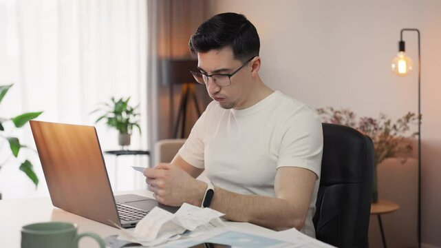 Tax season struggles as a tired man sorts through bills and checks, working on his laptop in a warm, cozy home interior. Financial responsibility meets exhaustion in a remote setting