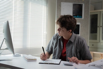 Young man focused on analyzing data at a desk, using a computer and tablet. Sunlight filters through blinds, creating a productive environment. Office setting suggests a tech or design profession