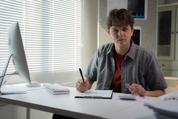 Fototapeta premium Young professional sitting at a desk in a modern office, analyzing data on a tablet. Bright natural light streams through blinds, creating a focused and productive atmosphere