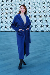 Elegant woman in a stylish blue coat standing against a modern architectural background. She smiles confidently, exuding warmth and sophistication. Outdoor portrait with natural lighting.