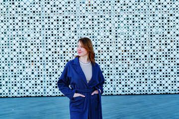 Elegant woman in a stylish blue coat standing against a modern architectural background. She smiles confidently, exuding warmth and sophistication. Outdoor portrait with natural lighting.