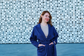 Elegant woman in a stylish blue coat standing against a modern architectural background. She smiles confidently, exuding warmth and sophistication. Outdoor portrait with natural lighting.