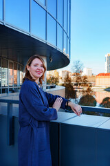 A stylish young woman in a blue coat stands outside a modern glass building, leaning on a railing and smiling. The reflective windows of the structure capture the surrounding cityscape.