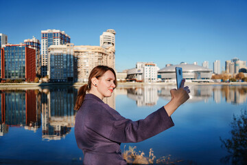 Young woman taking a selfie with a smartphone against a modern cityscape and water reflections. Concept of urban lifestyle, tourism, social media, and digital connection.