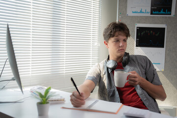 Young man sits at a desk with headphones around his neck, holding a coffee mug and writing in a...