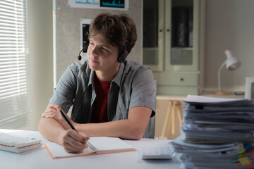 Young man wearing a headset, focusing on studying at a desk filled with papers and notebooks. Bright natural light filters through blinds, creating a productive home office atmosphere