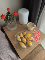 Valentine's day breakfast with fresh croissants, coffee, red roses, and a heart patterned napkin on a bedside table