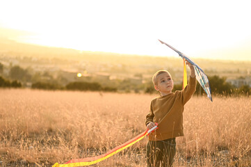 A boy holds a kite in his hands, standing in a field during sunset. The warm golden light casts a magical glow over the scene, capturing the joy of childhood and outdoor adventure.