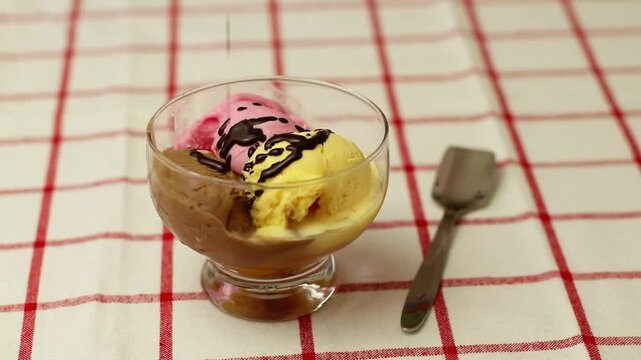 chocolate goteando sobre una copa de helado sobre un mantel blanco con lineas rojas y una cuchara