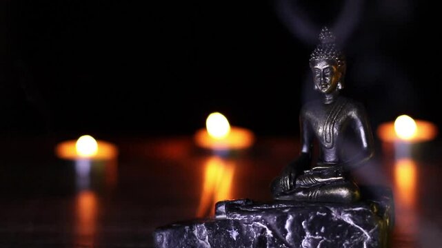 buda sentado sobre piedra en primer plano, al fondo luz c&aacute;lida de velas encendidas y humo de incienso