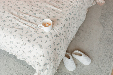 Overhead view of a cup of coffee on a bed with a floral duvet and a pair of slippers