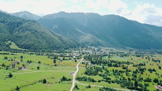 White clouds gently caress towering mountain peak, casting shadows on bright green valley below, where dirt road winds through picturesque landscape. Shooting from drone.